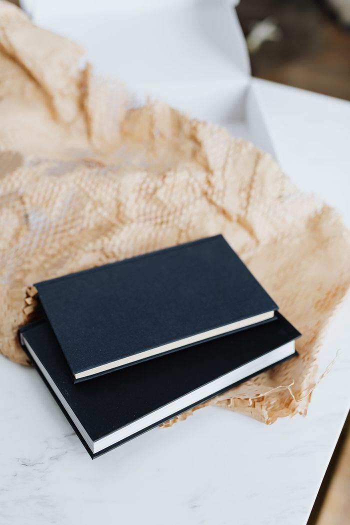From above of notebook pair with black empty hardcover placed with packing paper and white cardboard box on white marble table